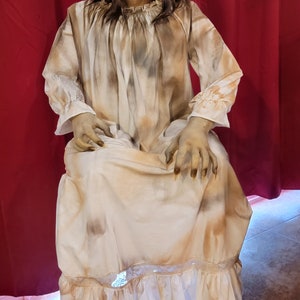 May include: A seated Halloween decoration of a pale-skinned, aged figure with long, dark hair. The figure wears a stained, cream-colored dress with long sleeves and a ruffled hem. The figure has long, sharp fingernails and a menacing expression.