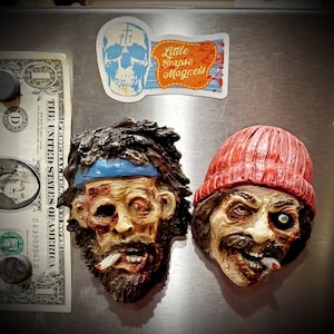 May include: Two refrigerator magnets depicting zombie-like faces. The first is a man with a blue bandana and a cigarette hanging from his mouth. The second is a man wearing a red beanie and also has a cigarette hanging from his mouth. The magnets are on a silver refrigerator.
