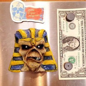 Könnte beinhalten: Ein gelber und blauer Keramikmagnet, der ein Comic-Gesicht mit einer Pharaonenkopfbedeckung zeigt. Der Magnet befindet sich an einem silbernen Kühlschrank neben einem Dollar-Schein und einem weiteren Magneten. Der Dollar-Schein trägt den Text "THE UNITED STATES OF AMERICA" und "ONE DOLLAR".