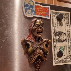 May include: A zombie bust magnet with green, brown, and red tones, featuring exposed flesh and a decaying appearance. A skull magnet and a dollar bill are also attached to the metal surface. The magnet is a unique piece of horror-themed decor.