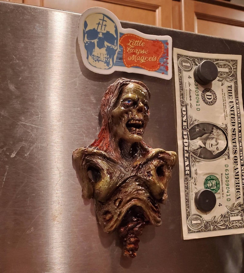 May include: A zombie bust magnet with green, brown, and red tones, featuring exposed flesh and a decaying appearance. A skull magnet and a dollar bill are also attached to the metal surface. The magnet is a unique piece of horror-themed decor.
