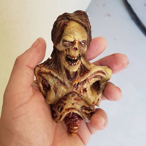 May include: A detailed, hand-painted zombie bust with exposed flesh and a gruesome appearance. The figure has a decaying complexion, open mouth, and detailed hair. The bust is held in a hand.