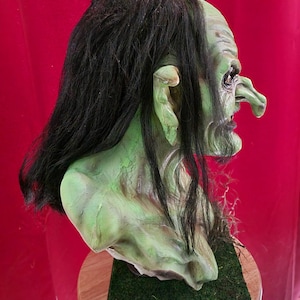 May include: A green and brown goblin bust with long black hair. The bust is on a green grass-like surface.