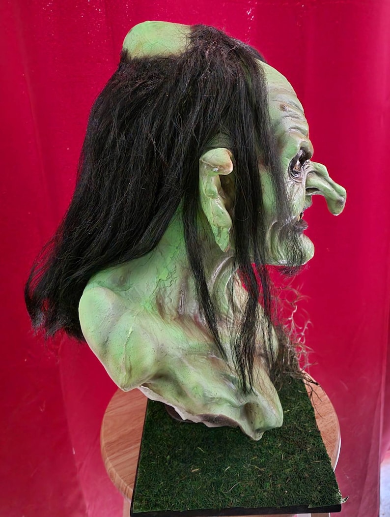 May include: A green and brown goblin bust with long black hair. The bust is on a green grass-like surface.