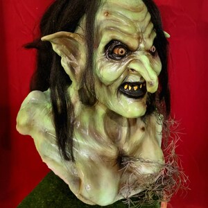 May include: A green goblin mask with long black hair, pointed ears, and a large, crooked nose. The mask has a menacing expression with sharp teeth and piercing yellow eyes.