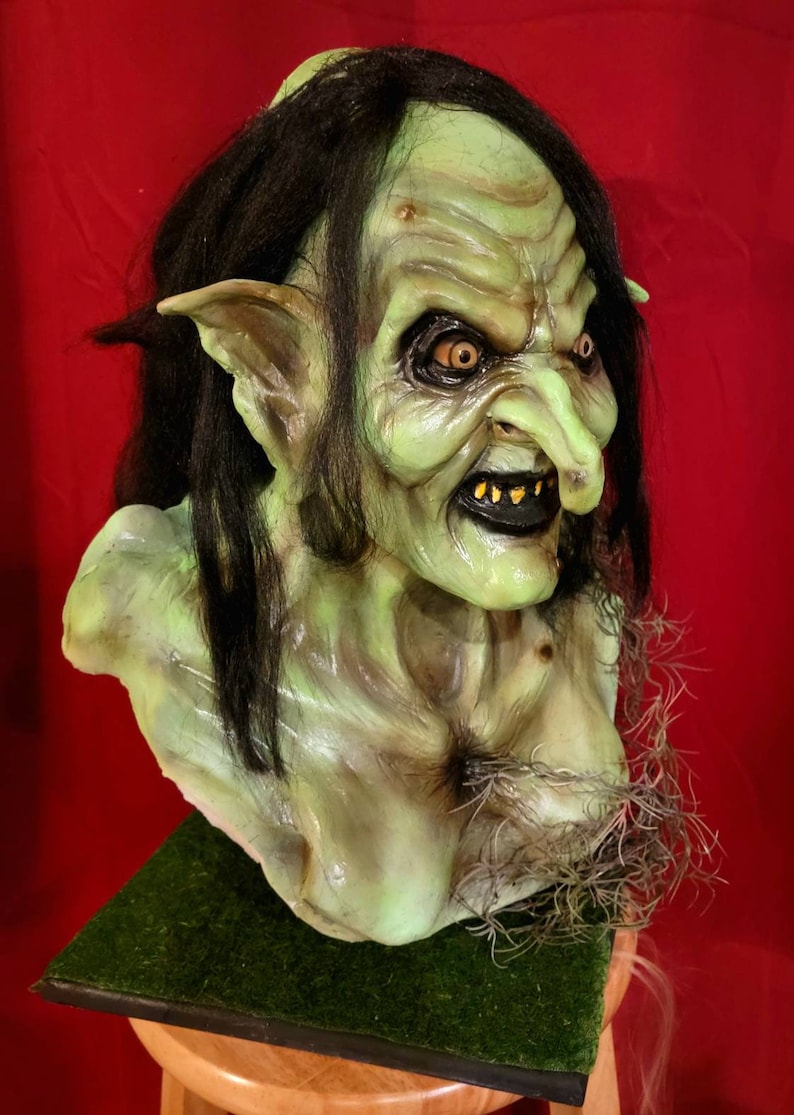 May include: A green goblin mask with long black hair, pointed ears, and a large, crooked nose. The mask has a menacing expression with sharp teeth and piercing yellow eyes.