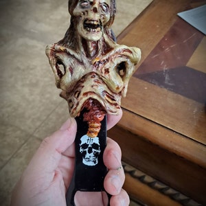 May include: A zombie-themed bottle opener. The opener features a detailed, decaying zombie bust in shades of beige, brown, and red. The black metal opener has a skull and crossbones design with the text "&dagger;&dagger;".
