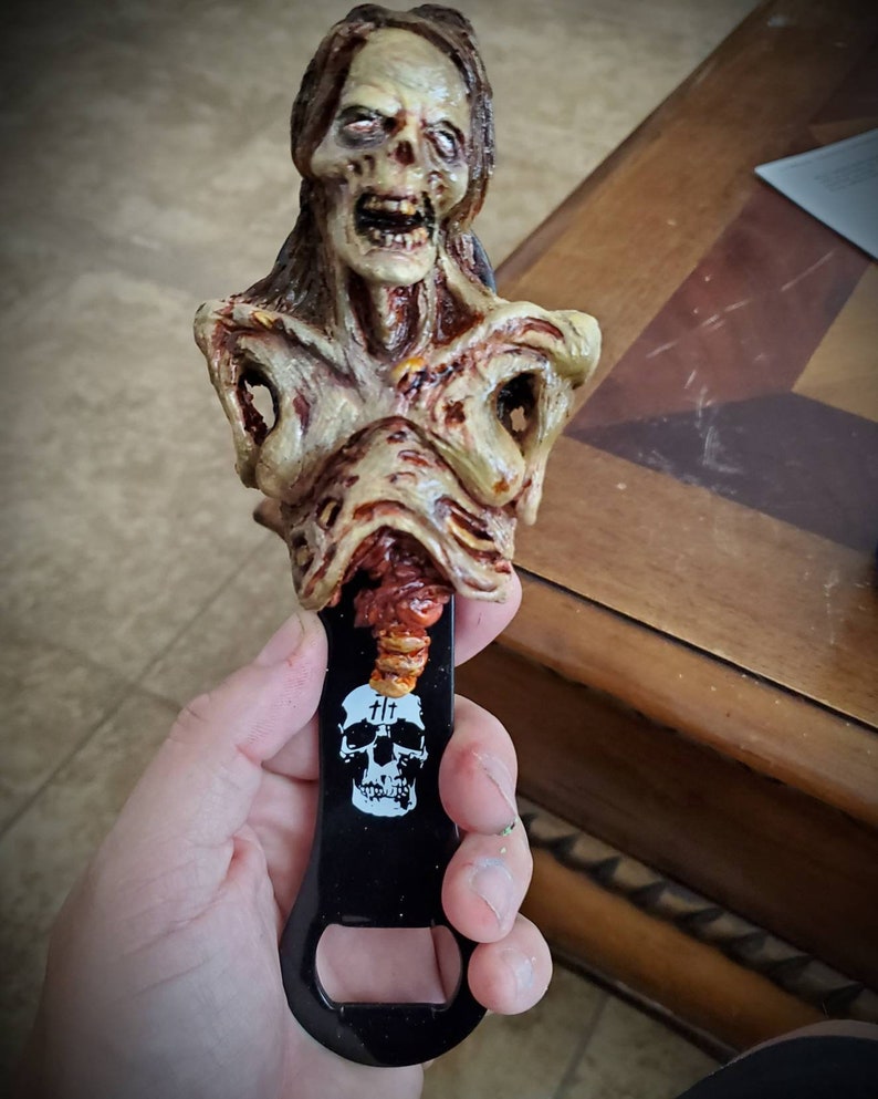 May include: A zombie-themed bottle opener. The opener features a detailed, decaying zombie bust in shades of beige, brown, and red. The black metal opener has a skull and crossbones design with the text "&dagger;&dagger;".