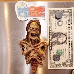 May include: A zombie-themed magnet with a detailed, decaying appearance. The magnet is tan, brown, and red, with visible ribs and exposed flesh. A one-dollar bill and two magnets are next to the zombie magnet. The text "Little Corpse Magnets" is on a sticker.
