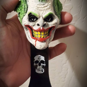 May include: A bottle opener featuring a sculpted Joker head with green hair, white face, and a red and yellow grin. The black handle has a skull graphic. A unique, novelty bar accessory.