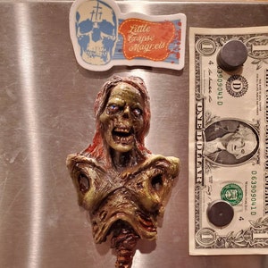May include: A zombie-themed magnet with a detailed, decaying bust. The magnet is green, brown, and red, with blue eyes. Above it are two stickers: one with a skull and crosses, and another with the text "Little Corpse Magnets".