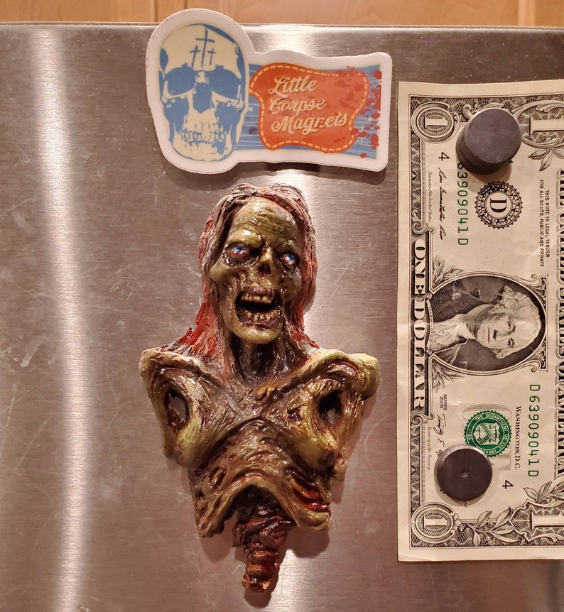 May include: A zombie-themed magnet with a detailed, decaying bust. The magnet is green, brown, and red, with blue eyes. Above it are two stickers: one with a skull and crosses, and another with the text "Little Corpse Magnets".
