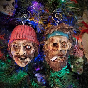 May include: Two Christmas ornaments shaped like human heads. One is wearing a red knit hat and the other has a blue headband. Both have realistic facial features and are decorated with details like a cigarette and a beard.