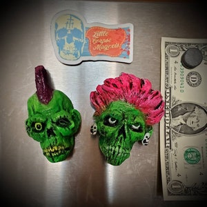 May include: Green zombie skull magnets with pink and purple mohawks and a sticker that says "Little Corpse Magnets". A dollar bill is next to the magnets.