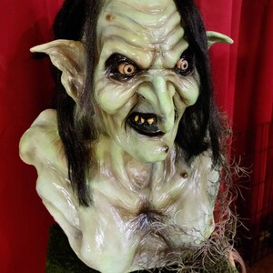 May include: A green, grotesque, goblin-like bust with long black hair, pointed ears, and a large, bulbous nose. The bust is made of a realistic-looking material and has a menacing expression.