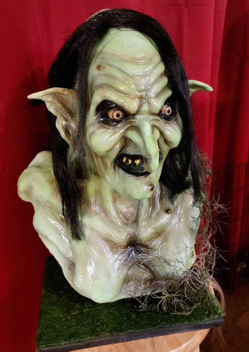 May include: A green, grotesque, goblin-like bust with long black hair, pointed ears, and a large, bulbous nose. The bust is made of a realistic-looking material and has a menacing expression.