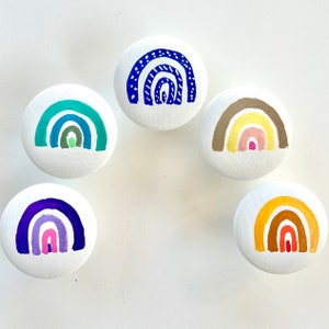 Peut inclure: Six boutons ronds en céramique blanche avec des motifs arc-en-ciel peints. Chaque bouton présente un schéma de couleurs et un motif d'arc-en-ciel différents.