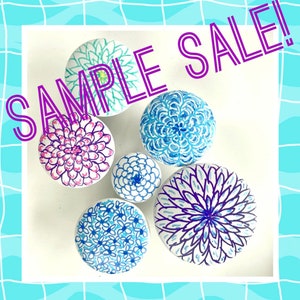May include: Six round ceramic knobs with hand-painted floral designs in shades of blue, pink, and purple. The knobs are arranged in a circle on a white background. The text "SAMPLE SALE!" is written in purple letters across the top of the image.