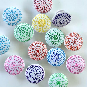 May include: Twelve round white ceramic knobs with colorful mandala designs painted on them. The designs are in shades of red, orange, yellow, green, blue, purple, and pink.