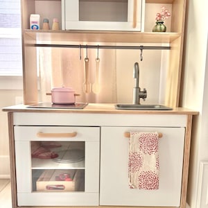 Peut inclure: Une cuisine jouet en bois avec un micro-ondes blanc, une casserole rose sur une plaque de cuisson, un évier argenté avec un robinet et des ustensiles en bois suspendus. Le bas comprend un four vitré et un placard avec un torchon à motifs floraux.