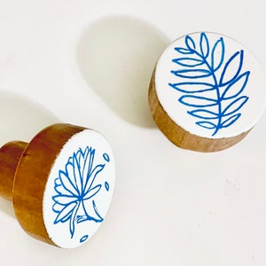 Könnte beinhalten: Zwei Holzknöpfe mit blauen und weißen Blumenmustern. Die Knöpfe sind rund und haben einen weißen Hintergrund mit blauen Blumenmustern.