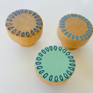 May include: Three wooden knobs with a decorative blue and black border. The knobs are round and have a light brown color. One knob has a light green center.