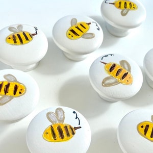 Pode incluir: Conjunto de 8 botões de gaveta de cerâmica branca com desenhos de abelhas pintados à mão. Cada botão apresenta um desenho de abelha diferente com listras amarelas e pretas.