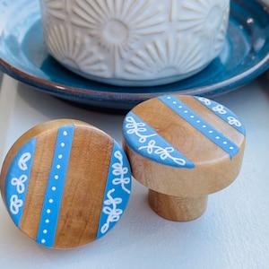 May include: Two wooden knobs with a blue and white painted design. The knobs have a light brown wood base and are painted with a blue stripe and white dots. The knobs are decorated with white painted leaves.