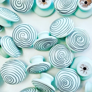 May include: A collection of 15 light blue cabinet knobs with black spiral designs. The knobs are made of wood and have a smooth, rounded surface.
