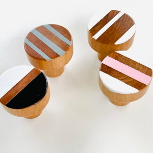 May include: Four wooden knobs with different painted stripes. The knobs are round and have a brown wood base. The stripes are painted in white, black, pink, and gray.