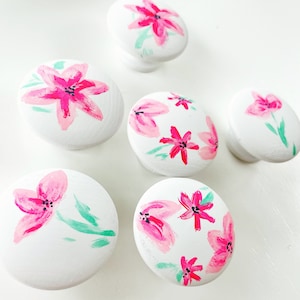 Pode incluir: Seis botões redondos brancos com desenhos florais cor-de-rosa pintados sobre eles. Os botões têm um diâmetro de aproximadamente 5 cm.