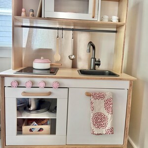 May include: A wooden play kitchen featuring a white microwave, a black stovetop, a sink with a faucet, and four pink knobs. The unit includes an oven with a glass door, a lower cabinet with a patterned towel, and hanging utensils on a black rod.