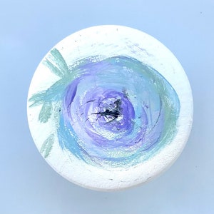 Pode incluir: Um descanso de copo redondo de cerâmica branca com um desenho pintado de uma flor roxa e azul com folhas verdes.