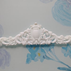 May include: White decorative architectural moulding with an ornate, symmetrical design. The moulding features a central oval shape and swirling, scroll-like details. The piece is made of a solid white material, likely for interior design or craft projects.