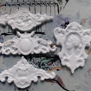 Könnte beinhalten: Vier weiße dekorative Verzierungen aus Kunstharz mit aufwendigen, Vintage-Designs. Die Formen umfassen ein Cameo, ein Wappen und zwei symmetrische Verzierungen, ideal für Bastelprojekte oder Heimdekoration.