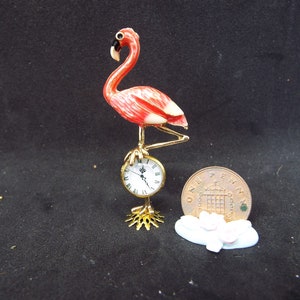 May include: A pink flamingo figurine standing on a gold clock with Roman numerals. The figurine is approximately 5 cm tall and is made of ceramic. The clock is approximately 2.5 cm in diameter. The figurine is on a white base with pink flowers.
