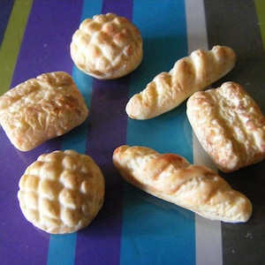 May include: A selection of six different types of bread rolls, including a round roll with a swirl pattern, a long loaf, and a few smaller rolls. The rolls are all a light brown colour and appear to be freshly baked.