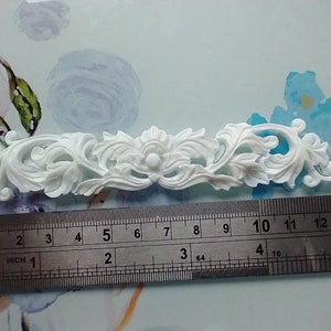 May include: A white, decorative floral and leaf design. The piece is approximately 13 cm long, measured with a ruler. The design features a central floral element with symmetrical, swirling leaf patterns extending outwards.