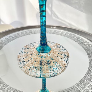 Op de afbeelding: Een decoratief teal en goudkleurig wijnglas met ingewikkelde handgeschilderde details. Het glas heeft een lange steel met gouden accenten en een brede, ronde basis versierd met gouden bladpatronen. Het glas staat op een spiegeloppervlak.
