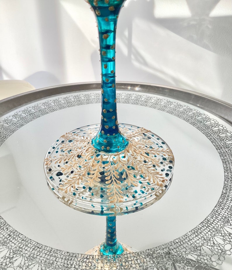 Op de afbeelding: Een decoratief teal en goudkleurig wijnglas met ingewikkelde handgeschilderde details. Het glas heeft een lange steel met gouden accenten en een brede, ronde basis versierd met gouden bladpatronen. Het glas staat op een spiegeloppervlak.