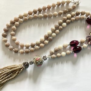 Könnte beinhalten: Eine lange, doppelfädige Halskette mit einem Quastenanhänger. Die Halskette besteht aus hellbraunen und weißen Perlen, mit violetten Glasperlen und silbernen Akzenten. Die Quaste besteht aus kleinen, hellbraunen Perlen.