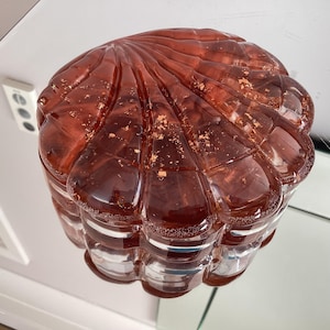 May include: A decorative, glass container with a lid. The container is a deep reddish-brown color with gold flecks and a ribbed, shell-like design. The glass is transparent, allowing light to pass through.
