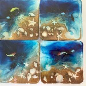 May include: Four square resin coasters with an ocean theme. Each coaster features a blue and gold design with embedded seashells, starfish, and small fish. The coasters have a glossy finish and are suitable for home decor.