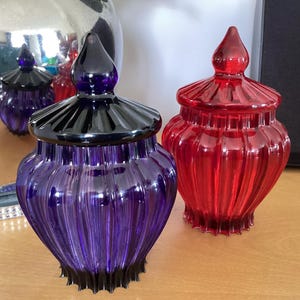 May include: Two decorative glass jars with lids. One is a deep purple with a black lid, and the other is a vibrant red. Both jars have a ribbed design and a pointed finial on the lid. They sit on a wooden surface.