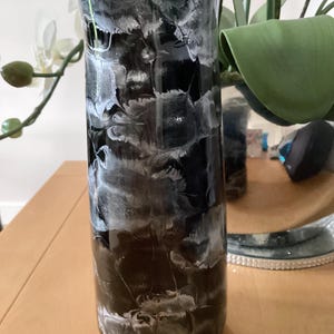 Könnte beinhalten: Eine hohe, zylindrische Vase mit einem schwarz-weißen Marmormuster. Die Vase hat eine glänzende Oberfläche und besteht aus Glas. Das Design ähnelt wirbelnden Wolken oder Rauch. Die Vase eignet sich zum Präsentieren von Blumen oder als dekoratives Element.