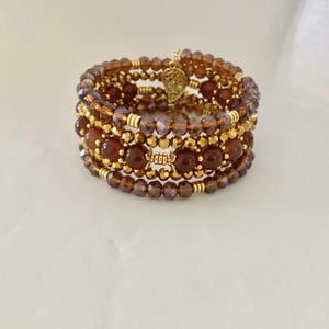 May include: A multi-strand wrap bracelet featuring translucent brown faceted beads, smooth round amber-colored beads, and small metallic gold-tone spacer beads. The bracelet includes a gold-tone metal leaf-shaped charm attached to the top strand.