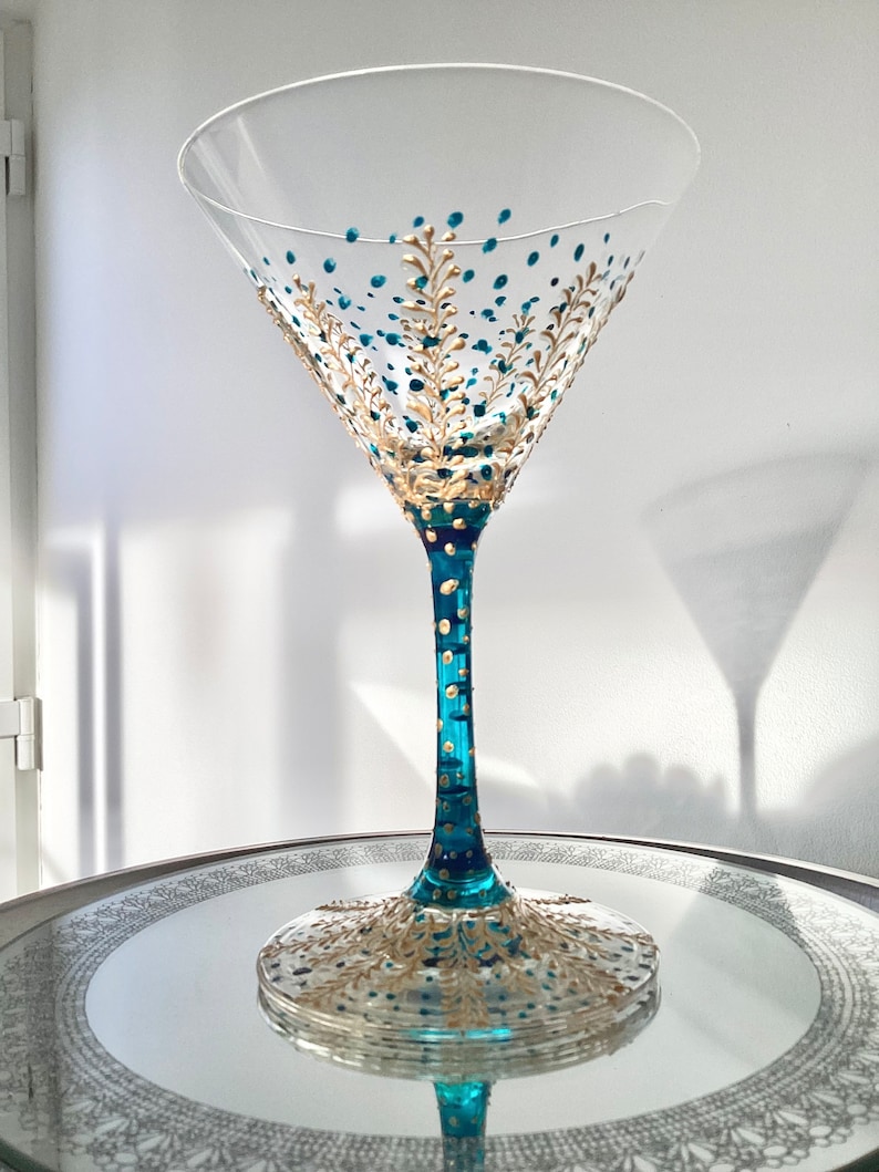 Op de afbeelding: Een helder martini glas met een teal en goud geschilderd ontwerp. Het glas heeft teal stippen en gouden bladpatronen op de kom en voet. De steel is teal met gouden accenten. Het staat op een zilveren dienblad.