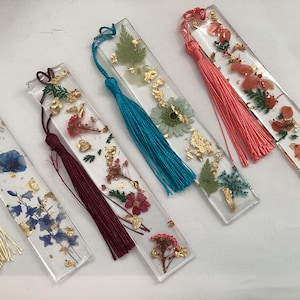 Puede incluir: Marcapáginas rectangulares de resina transparente con flores secas y detalles de pan de oro. Cada marcapáginas tiene una borla de diferente color, incluyendo dorado, burdeos, turquesa y coral. Los marcapáginas muestran varios arreglos florales.