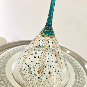 Op de afbeelding: Een handbeschilderd martiniglas met een turquoise steel en gouden details. De heldere glazen kom is versierd met gouden en turquoise blad- en stippatronen. Het staat op een zilveren dienblad met een decoratieve rand.