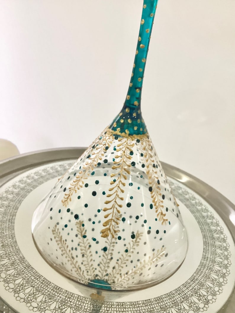 Op de afbeelding: Een handbeschilderd martiniglas met een turquoise steel en gouden details. De heldere glazen kom is versierd met gouden en turquoise blad- en stippatronen. Het staat op een zilveren dienblad met een decoratieve rand.
